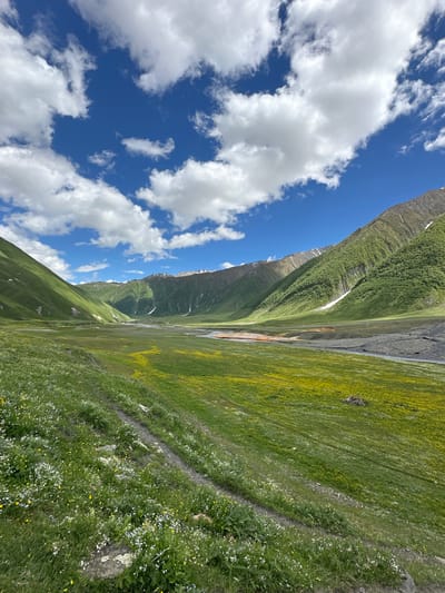 The image captures a scenic view of a valley in Ketrisi, Georgia, under a bright blue sky dotted with fluffy white clouds. Lush green rolling hills flank the valley, with patches of snow visible on the higher slopes and mountain peaks. A river, its water appearing reddish-orange in some sections, flows through the valley floor. The foreground is covered in vibrant green grass and dotted with small white and yellow wildflowers. A dirt path winds its way up the grassy slope on the left side of the image. The overall impression is one of natural beauty and tranquility.