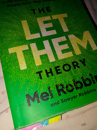 The image shows a close-up, angled shot of the cover of a book titled "The Let Them Theory" by Mel Robbins and Sawyer Robbins. The book cover is bright green with a pattern of small yellow dots. The title is in large, bold letters, with "THE" in black at the top, "LET THEM" in large, white letters with yellow outlines in the center, and "THEORY" in black below that. The authors' names, "Mel Robbins" and "and Sawyer Robbins," are also in black at the bottom of the cover. The book is lying on a textured fabric, possibly a blanket or tablecloth, with a pink polka dot pattern visible in the lower left and lower right corners. The lighting is dim, and there are some minor scuffs and dust marks on the book cover. The scene is a personal space, likely indoors, and there is no indication of it being located in the city of Cimahi, Indonesia.