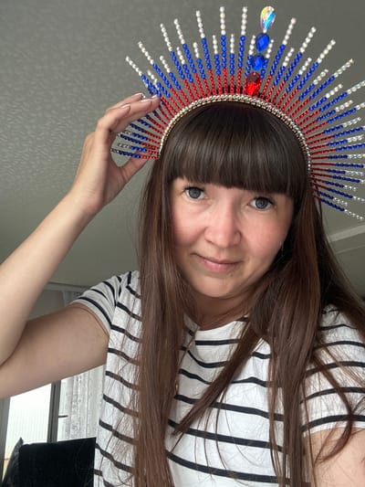 A woman with long brown hair and bangs is wearing a crown adorned with red, white, and blue rhinestones, with larger red and blue gems in the center. She is wearing a black and white striped t-shirt and is holding her hand to the side of the crown. The background is a plain gray wall. The image appears to have been taken indoors. The location of this image is not specified in the provided information.