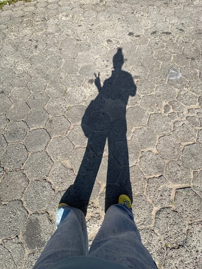 The image shows a downward view of a person standing on a hexagonal-patterned stone walkway. The person is wearing light blue jeans and bright yellow footwear with visible designs. The person's shadow is cast on the walkway, with one hand gesturing in a "peace" sign. A small, clear plastic bag is visible on the walkway to the left of the shadow. The location is in Florianópolis, Brazil.