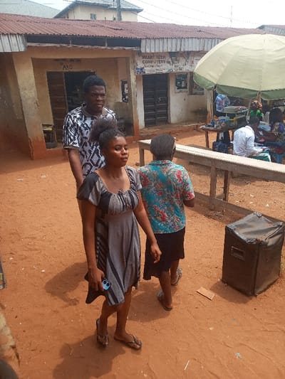 The image displays an outdoor commercial area in Nsugbe, Nigeria, under bright daylight with a reddish-brown earth ground.

In the foreground, three individuals are walking from left to right. A female in a grey and black patterned short-sleeved dress and black flip-flops holds a dark smartphone in her left hand, with her dark hair tied up. Behind her, a male wears a black and white patterned short-sleeved shirt and dark pants. To the right of the woman, a third individual, viewed from the back, wears a blue floral shirt, a black skirt, and dark flip-flops. To the far right, a black, rectangular, fabric-covered case or speaker rests on the ground.

The midground features a long, light-brown wooden bench. Behind the bench, a light-green and beige umbrella shades a market stall. Several individuals are present at the stall, including one person in a white shirt seated at a table displaying a printer, papers, and other items.

The background consists of a single-story building with a reddish-brown corrugated metal roof and light-brown walls. An open doorway is visible, marked "A30." Adjacent to it, a closed doorway with dark metal gates features a sign above it reading "SMART TECH COMPUTERS." Various posters and papers are affixed to the building's facade. A portion of another building with a grey roof is visible in the distant background.
