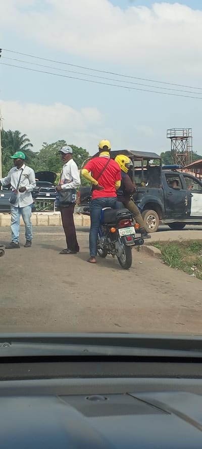 An outdoor scene in Akure, Nigeria, depicts a roadside interaction under a bright, cloudy sky. Four adult males are prominent. On the left, two dark-complexioned men stand by the road's edge. The leftmost wears a green cap, light shirt, and blue jeans, carrying a light blue sling bag. The man beside him wears a grey cap, white graphic shirt, and dark trousers, with a dark sling bag. To their right, a dark-colored TVS motorcycle, with Nigerian license plate KJK-558XC, is stopped. Its driver, seen from behind, wears a yellow cap, a red shirt over a yellow long-sleeved shirt, and blue jeans, with a black sling bag. A passenger, wearing a bright yellow full-face helmet, dark jacket, and dark trousers, sits behind him. Directly behind the motorcycle, a dark-colored police pick-up truck with white accents and a roof-mounted light bar is stopped. An individual in a light top is partially visible in the truck's driver's seat. In the background, a dark SUV with an open hood is parked, alongside other vehicles. Green foliage and a tall metal lattice structure are visible further back. The photograph is taken from inside a vehicle, with the dashboard visible at the bottom.