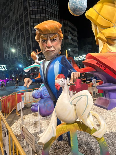 The image captures a nighttime street scene in Alicante, Spain, likely during the Fallas festival. In the foreground, a large, colorful statue, possibly a caricature of Donald Trump, dominates the frame. He is depicted with a stern expression, wearing a blue suit. To his right, a white swan-like figure with its eyes closed and a red cap on its head is positioned. Behind the statue, palm trees and illuminated buildings line the street. The ground is covered in gravel. A yellow barrier is visible in the foreground, with several informational signs displayed. The atmosphere suggests a public event with artistic installations.