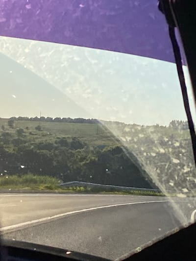 A road perspective through a dirty car window shows a rural landscape. The foreground is dominated by the asphalt of a road with a white line marking. Beyond the road, rolling hills covered in green vegetation and trees are visible. In the distance, a treeline with scattered structures and what appear to be power lines stretches across the horizon under a bright, hazy sky. The view is partially obscured by the purple tint of the car's top window and smudges or debris on the glass. The scene suggests a journey through a rural area, potentially near Заглядовка, Russia, given the context, under daylight conditions.