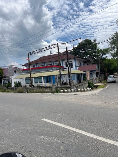 The image captures a street view in the city of Langsa, Indonesia, focusing on a building complex. The building, painted in white and blue with red accents, features a prominent red-tiled roof. In front of the building, there is a low, decorative stone fence. A metal truss structure extends over a portion of the building. A sign is visible on the left side of the building, with text that appears to read "UPTD LABORA. ORUM KESEHATAN KOTA LANGSA". The road in the foreground has a white line marking. A silver sedan is parked on the right side of the road. Trees and a cloudy sky are present in the background.