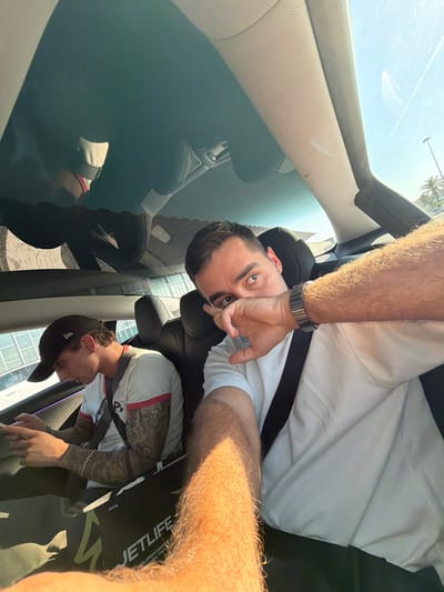 In this selfie, taken from a car's interior, two young men are the subjects. One man, wearing a white t-shirt and a watch, is positioned in the foreground, looking directly at the camera with his hand near his mouth. His arm, covered in hair, extends towards the top of the frame. The second man, wearing a white t-shirt with red trim and a black baseball cap, is seated in the back. He has tattoos on his arms and is looking downwards. The car's interior, with black seats and a light-colored headliner, is visible. The car appears to have a sunroof, showing a bright sky with palm trees, suggesting it is daytime with clear weather. Reflections on the car's surface indicate an urban environment with modern buildings. The overall mood seems casual and relaxed. The text "JETLIFE" is visible on the second man's shorts. The location context is el Prat de Llobregat, Spain.