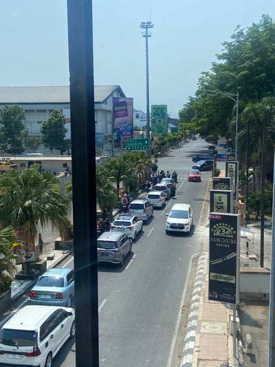 The image shows a street scene in Kudus, Indonesia, on a bright, sunny day. A multi-lane road is filled with various vehicles, including cars and motorcycles, indicating moderate traffic. On the right side of the road, there are tall palm trees and several vertical billboards advertising "234 MAGNUM KRETEK" with prices. The billboards are black with yellow text and graphics. Further down the road, there are more billboards and streetlights. On the left side of the road, there is a building with a large white roof, possibly a commercial establishment or factory, and some smaller buildings. Green trees and vegetation are visible throughout the scene. The sky is clear and blue. A large, dark vertical pole is prominently in the foreground, partially obscuring the view of the street.