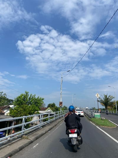The image shows a person riding a motorcycle on a bridge in Kecamatan Tanggulangin, Indonesia. The person is wearing a blue helmet and is driving the motorcycle with a black license plate that reads "W 6995 VP." The bridge is surrounded by a silver metal guard rail. The road is asphalt and a white car is driving in the opposite direction. The sky is clear and blue with some white clouds. There is a street sign on the right side of the image. The bridge appears to be in a suburban or rural area with houses and trees visible in the background.