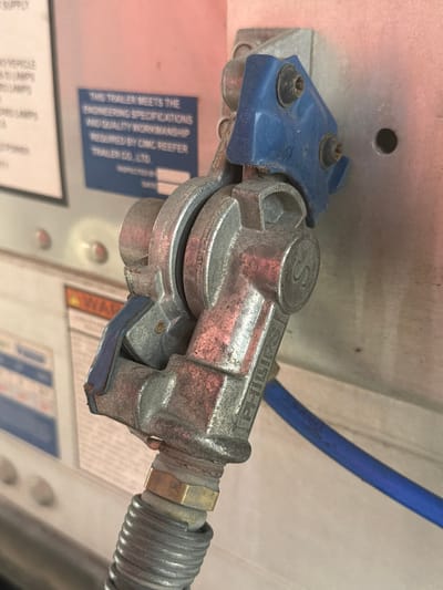 The image shows a close-up of a metal coupling with a blue plastic cap, attached to a gray corrugated hose. The coupling has the word "PHILLIPS" embossed on it. In the background, there are labels on a light-colored surface. The scene appears to be an outdoor industrial or automotive setting, possibly at a truck stop or repair facility. The lighting suggests it is daytime. There is no indication of the city of Murfreesboro, United States of America, nor any people or specific actions.