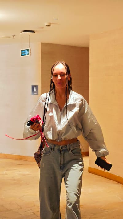 A woman with long, dark hair styled in numerous small braids is depicted walking through an indoor corridor. She wears a light blue or grey cropped windbreaker jacket with a zippered front, exposing a sliver of her midriff, paired with light-wash, wide-leg jeans. In her right hand, she carries a red ribbon or strap along with a piece of red and black fabric, possibly a face mask or small item. A black smartphone is held in her left hand.
The setting appears to be a well-lit hallway or lobby, characterized by warm, beige-colored walls and a matching tiled floor. The white ceiling features several fixtures, including a light, a smoke detector, and another electronic device. A green illuminated "EXIT" sign, displaying an arrow pointing right and text in both English and what appears to be another language, hangs from the ceiling. Further along the wall, a "WOMEN" restroom sign, indicated by a female stick figure, is visible. The artificial lighting gives the space a bright, inviting atmosphere. The woman's expression is neutral, and she appears to be moving purposefully.