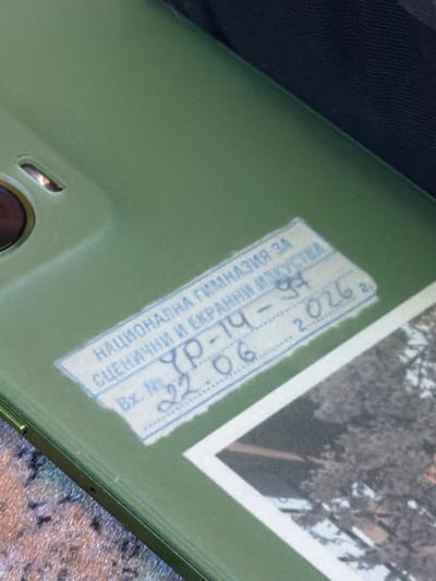 The image shows a close-up of the back of a light green object, possibly a phone or tablet case, with a textured dark fabric in the upper left corner and a patterned surface in the lower right. A white label with blue text is affixed to the object, diagonally across the center. The label is partially obscured by the object's surface and appears to be a stamp or a sticker with official information.

The visible text on the label is in Bulgarian and reads: "НАЦИОНАЛНА ГИМНАЗИЯ ЗА СЦЕНИЧНИ И ЕКРАННИ ИЗКУСТВА" which translates to "National High School for Stage and Screen Arts". Below this, there is an entry number "Bx. № YP-14-97" and a date "22.06.2026". The text indicates that this is a document related to the National High School for Stage and Screen Arts, with an entry number and a future date. The overall impression is of an official document or stamp applied to a personal item. The background appears to be a photograph or painting, possibly of trees with snow or frost, visible in the upper right quadrant, framed by a white border. The lighting suggests it is daytime.