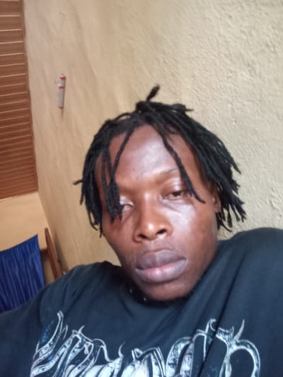 The scene depicts a close-up, head-and-shoulders view of an adult male with a dark complexion and black dreadlocks, appearing to be an indoor setting in what is reportedly Obwe, Nigeria. The individual wears a dark-colored t-shirt with a light-toned graphic design across the chest, and a thin, dark choker or necklace. His face is oriented directly towards the camera, with a neutral expression; the left eye appears partially closed or heavy-lidded, while the right eye is slightly more open. A light sheen is visible on the skin. The background consists primarily of a textured, light brown/tan wall. To the upper left, a red and white disposable cigarette lighter is vertically affixed to the wall. Further left, a section of dark blue, pleated fabric, likely a curtain, is visible, partially obscuring a vertical wooden structural element. No other individuals or direct interactions are depicted. The lighting is even, suggesting ambient indoor illumination.