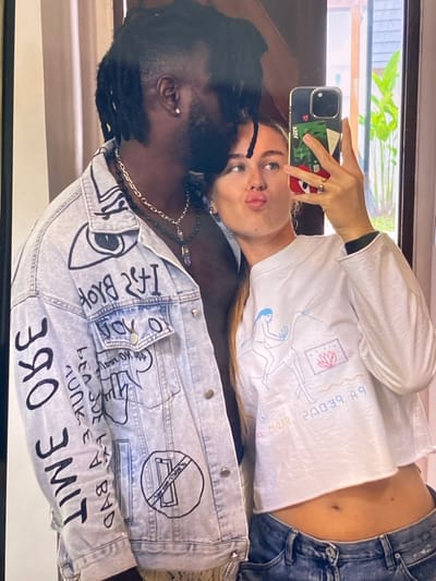 A dark-skinned man with dreadlocks and a woman with blonde hair are taking a selfie in a mirror. The man is wearing a denim jacket covered in black graffiti. The woman is wearing a white crop top and blue jeans. She is holding a phone and making a kissing face. The background shows a doorway with greenery visible outside. The image appears to be taken in a residential setting.