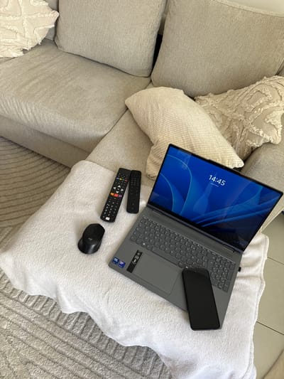 Here is a detailed description of the image:

**Scene Overview:**

The image depicts a relaxed, comfortable setting, likely indoors. A laptop is open on a large, soft white cushion, which rests on a beige colored couch covered with pillows.  Various other items are scattered around.

**Objects Present:**

*   **Laptop:** The laptop screen displays a blue background with the time "14:45" and date "jeudi 9 avril" (Thursday, April 9th) visible in the center.
*   **Smartphone:** A black smartphone lies atop the laptop's keyboard area.
*   **Remote Controls:** Two remote controls, one black and one with colorful buttons, are placed near the laptop.
*   **Mouse:** A black Logitech computer mouse sits to the right of the remote controls.
*   **Couch:** The laptop sits on a white cushion on a large couch with several beige pillows.
*   **Pillows:** The couch contains multiple pillows, including some with a decorative white trim.

**Environment:**

The environment appears to be a comfortable living area. The lighting is natural and soft, suggesting daytime. The overall ambiance implies relaxation and leisure.

**Location:**

The displayed date "jeudi 9 avril" indicates a European setting, but there is no information about the city of Cannes, France from the image.