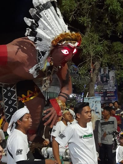 An outdoor nighttime scene in Kedungbanteng, Indonesia, features a large, anthropomorphic float dominating the frame. The float depicts a muscular figure with reddish-brown skin, adorned with a substantial white and black feathered headdress and a reddish-brown fur-like headband. Its face presents a grimacing expression with intensely glowing red eyes. A red band marks the left bicep, and black tribal patterns are visible on the arm. The lower body of the float is partially covered by a garment featuring white and black geometric designs.

In the immediate foreground, an adult male with a white cloth headwrap, a white short-sleeved t-shirt with a dark "JG" or "IG" logo, and a black long-sleeved undershirt stands facing left. To his right, another adult male wears a white short-sleeved t-shirt with a green and black "INDAH" or "INTAN" logo, looking slightly right. Both individuals appear to be participating in an organized event.

A diverse crowd of people is visible in the background, indicating a public gathering. The environment includes a leafy tree on the right side and several banners/posters, one prominent blue banner displaying "CARNAVAL 2023", "MUDA ROLLAS", and "PRO AUDIO" text and logos. Artificial lighting illuminates the scene, with the float's glowing eyes providing a significant light source.