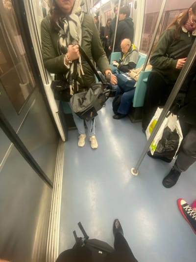 A passenger-eye-level view inside a subway car. Passengers are seated and standing, holding onto a horizontal metal pole. Bags, purses, and various footwear are visible. Seating is turquoise. One person wears red and white sneakers, another black dress shoes. Walls are white and gray.