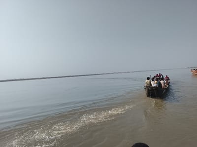 A wide, murky river in Ibi, Nigeria, under a hazy, bright sky, is depicted. In the midground, a dark wooden longboat transports approximately 10-15 individuals, moving away from the viewer and generating a visible wake. Passengers are seated, attired in light-colored clothing; some wear head coverings. A thin, flat landmass marks the distant horizon. A portion of a second wooden boat is visible on the far right. The immediate foreground water exhibits significant surface disturbance.
