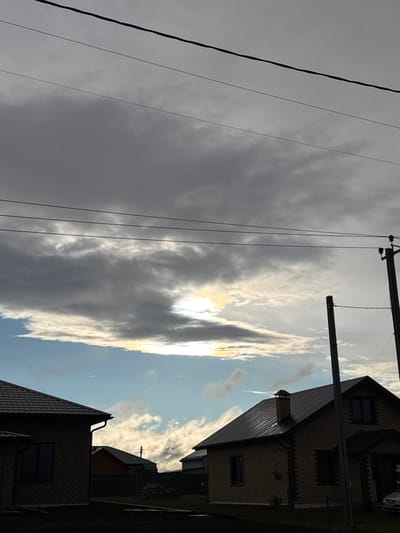The image depicts an outdoor scene in Gornyi (Dvurechenskoe S/P), Russia. Power lines run diagonally across the left side of the frame against a backdrop of grey, overcast skies. A column of sunlit clouds stretches vertically towards a blue sky. Portions of two brick buildings with dark-tiled roofs are visible on the right side of the image.