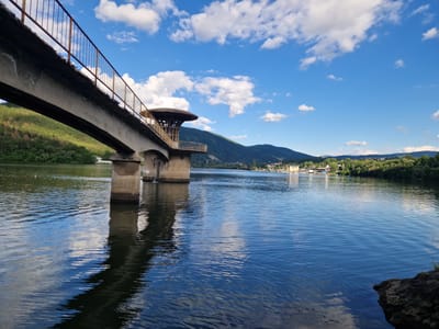 The image depicts a picturesque scene at the Pancharevo Lake in Pancharevo, Bulgaria. A concrete bridge with a rusty railing extends across the frame from the left foreground, supported by substantial concrete pillars. One of these pillars is visible in the water, casting a reflection on the rippling surface. The bridge leads to a circular, tower-like structure with a wooden roof. Beyond the bridge, the lake stretches into the distance, reflecting the bright blue sky dotted with fluffy white clouds. The far bank of the lake is lined with lush green trees, and a town or settlement with buildings and more greenery can be seen nestled against rolling, tree-covered hills in the background. The overall atmosphere is serene and summery, with the clear water and vibrant blue sky suggesting good weather. There are no people visible in the image.