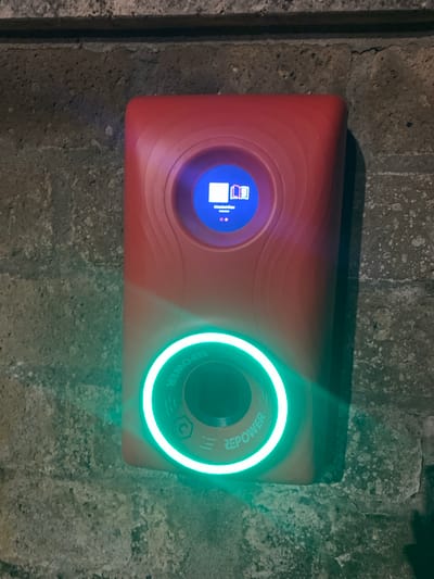 The image shows a red charging station mounted on a textured, grey wall, likely concrete or stone. The charging station has two circular interfaces. The left interface has a blue screen displaying an icon resembling an open book and some text, which is too blurry to read. The right interface is illuminated with a bright green light that forms a circular ring around a dark circular area. The text "REPOWER" is visible around the outer edge of this illuminated circle. Small graphic symbols, including what appears to be a crescent moon and three vertical lines, are also present within the illuminated circle. The overall lighting suggests it might be evening or in a dimly lit indoor space, given the artificial glow from the charging station. The texture of the wall and its rough appearance might indicate an outdoor or industrial setting. There are no people visible in the image.