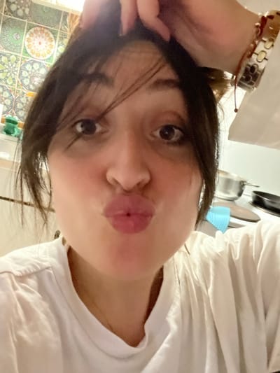 The image is a close-up, angled selfie of a person, likely female, making a "kissing" face. The person's head is tilted, with their face occupying the central and lower-right portion of the frame. They have dark, straight hair, some strands of which fall across their forehead and cheeks. Their eyes are dark and appear to be looking towards the camera. The lips are puckered into a distinct "kissing" or "pout" shape, with a reddish-pink color. The skin tone is fair.

On the upper left side of the image, part of the person's wrist and forearm are visible, adorned with a multi-strand bracelet featuring light-colored, possibly metallic or shell-like, rectangular or oval beads, along with some reddish cord elements.

The person is wearing a white t-shirt or top, visible on the right side of the image, showing some fabric texture around the neckline area.

The background is somewhat out of focus but provides contextual clues. On the lower left side, a patterned tiled wall is visible. The tiles feature intricate, possibly Moroccan or mosaic-style, designs in shades of green, brown, and off-white, with circular and geometric motifs. Above and to the right, behind the person's head and shoulder, hints of kitchenware can be seen, including what appears to be a light blue colander or basket and metallic pots or pans, suggesting an indoor setting, possibly a kitchen.

The lighting appears bright and even, likely from an indoor light source, casting soft illumination on the person's face. There is no visible text in the image. The overall impression is one of a casual, playful moment captured in a personal photograph.