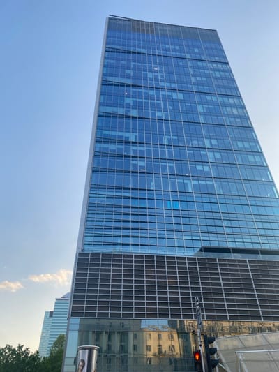 The image shows a tall, modern office building with a glass facade. The building is located at the coordinates 52.235, 21.009, which is in the city of Warszawa, Poland. The building is reflected in the windows of the building below, which is also a modern office building with a glass facade. There are several other buildings in the background, including a taller building to the left of the main subject. The sky is blue with a few clouds, and the sun is shining brightly. The image is taken from a low angle, looking up at the building. There is a traffic light on the right side of the image, with a red light illuminated. The image is likely taken from a street or sidewalk, as there is a small section of a sidewalk visible in the foreground. There is a small section of a bus stop in the bottom right corner of the image. There are no people visible in the image.
