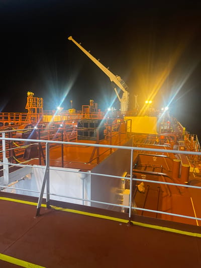A night scene shows the deck of a large tanker ship with a reddish-brown hull. Bright lights illuminate various structures, including an extendable white crane. Metal railings line the deck. Orange and yellow structures are also visible, possibly related to loading or unloading operations. The surrounding environment is dark.