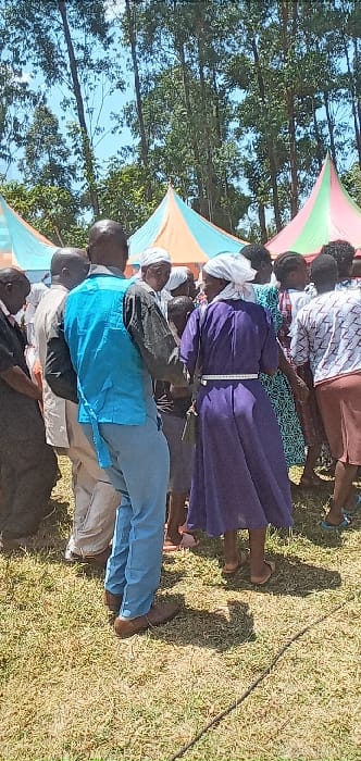 This image captures a group of people gathered outdoors in Burundu, Kenya, under bright, sunny conditions, likely midday or early afternoon given the prominent shadows on the grass. The scene features several adults, both men and women, attending what appears to be a community or social event.
In the background, two colorful tarpaulin structures, serving as shade covers or temporary shelters, are visible. One is pink and green, and another is light blue and orange. Tall trees with green foliage line the horizon, indicating an open, natural setting.
The people are positioned in what seems to be rows, mostly facing away from the camera or in profile. Notable attire includes a woman in the midground wearing a long, deep purple dress with a white head covering, and a measuring tape visible around her waist. In front of her, a man wears a light blue shirt over light-colored trousers. Other individuals are dressed in various casual and traditional clothing.
The ground is covered in dry-looking green grass, and strong shadows cast by the people indicate intense sunlight. A long, dark, thin object, possibly a cable or rope, lies on the grass in the lower right portion of the frame. The overall atmosphere suggests a communal outdoor gathering. There is no clearly readable text visible within the image beyond the unreadable markings on the measuring tape.