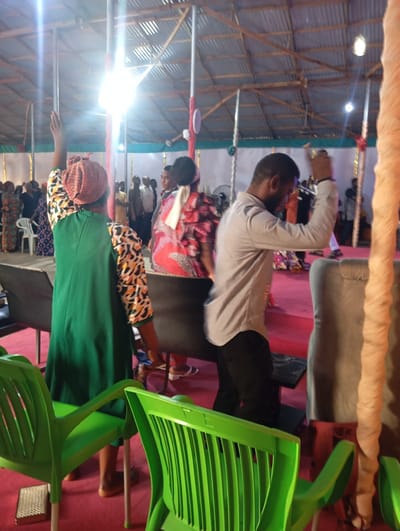 The image captures a gathering inside a building with a corrugated metal roof in Makurdi, Nigeria. The setting seems to be a religious meeting, possibly a church service. Several people are present, some standing and raising their hands in what could be a gesture of prayer or worship. Individuals are dressed in colorful traditional clothing. Chairs are arranged in the foreground, some being green plastic chairs, and others padded. A man in a light gray shirt and dark pants is standing and raising his hand in the middle of the room. The floor is covered with a red carpet. There are vertical poles decorated with fabric, possibly for a celebration. The scene is brightly lit, with some glare from the lighting fixtures.