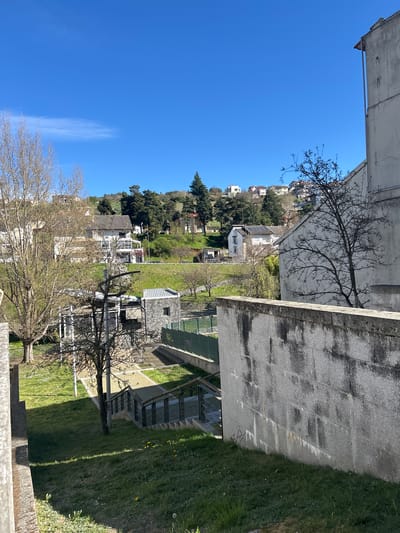 This image presents an elevated, static outdoor scene in Bragança, Portugal, capturing a residential and green landscape under a bright, clear blue sky with a single faint wispy cloud in the upper left. The immediate foreground on the right features a large, light grey concrete structure or wall, exhibiting prominent dark vertical staining from weathering, which casts a defined shadow onto a grassy slope. A bare-branched deciduous tree is positioned in front of and to the right of this wall. Below and to the left, a set of concrete stairs with dark metal railings descends into a lower-lying area of grass and paved pathways. Adjacent to the stairs and further left, a modern building with a grey stone facade and a metal roof is visible, situated next to a green fence that likely encloses a recreational court. The background reveals a hilly terrain ascending into the distance, characterized by a mix of bare and evergreen trees and multiple scattered residential houses. These houses are predominantly white or light-colored with visible tiled roofs. One specific house on the left side of the background features solar panels installed on its roof. No individuals, explicit actions, or interactions are discernible within the frame.