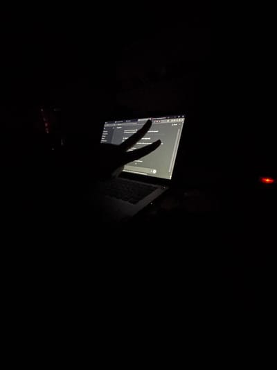 The image shows a dimly lit room with a laptop displaying a web page. A hand is gesturing in front of the screen, forming a "peace" or "victory" sign. The laptop is resting on a surface that appears to be a keyboard. Above the laptop is a red light, possibly from an electronic device. The overall environment is dark. There is no evidence to support the image being in Dobrovnitsa, Bulgaria.