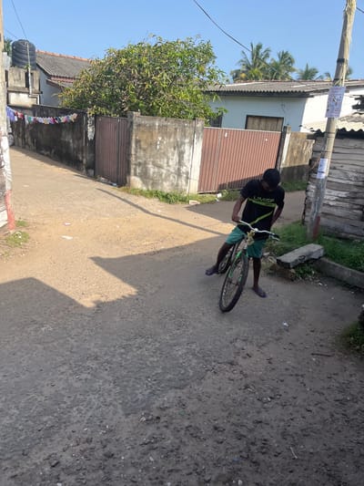 In Moratuwa, Sri Lanka, a young man in a black t-shirt and green shorts stands with a bicycle on a dusty, unpaved street. The bicycle is a mountain bike with a green frame and a white handlebar. The man is looking down at the bicycle. The street is lined with a weathered concrete wall on the left, on which laundry is hanging, and a wooden fence on the right. Behind the wall and fence, there are houses with tiled roofs and trees. The sky is clear and blue. The scene is illuminated by bright sunlight, casting shadows on the ground.