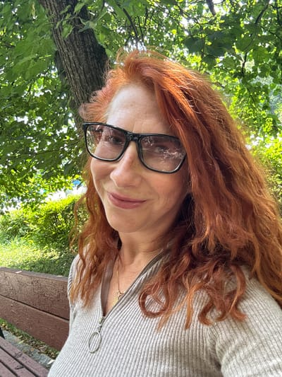 A woman with long, wavy, ginger hair and fair skin, wearing glasses and a grey ribbed sweater, is pictured outdoors. She is sitting on a wooden bench, with a tree and greenery in the background. The scene appears to be a park in Zelenograd, Russia.