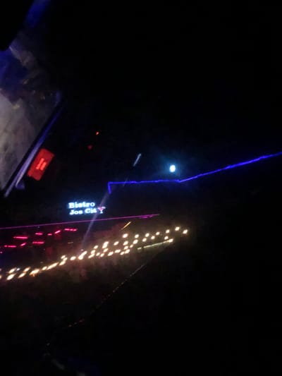 The image shows a nighttime scene with a focus on a lit sign that reads "Bistro Jos Cit" with a martini glass icon. Below the sign, there are rows of red and white lights, likely from a building or display. A blue neon-like light is visible in the upper right portion of the frame. The overall environment appears to be outdoors in a city, with the lights suggesting a commercial or entertainment area. The specific location of Rukuba Road, Nigeria, is mentioned in the prompt, but this is not visually evident in the image itself. There are no people visible in the image.