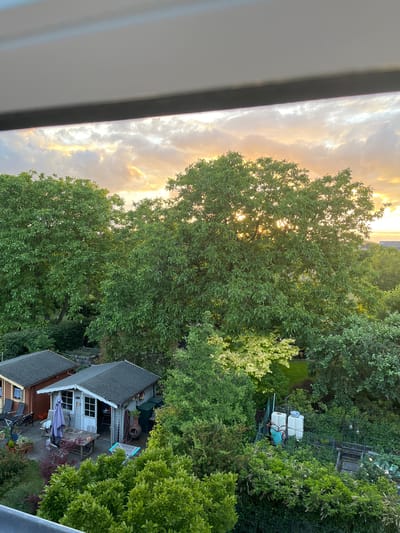 The image captures a tranquil backyard scene in Mondercange, Luxembourg, during what appears to be sunset or sunrise. Two garden sheds are visible in the foreground. The shed on the left is reddish-brown with a dark roof, and patio furniture, including chairs and a bench, is arranged outside. The shed on the right is painted light gray with a dark roof and features a wooden table with chairs and an umbrella set up on a patio area. Adjacent to the shed are two large white water tanks with some green piping.
Dominating the midground and background are lush green trees with dense foliage. The setting sun or rising sun's rays are filtering through the leaves of the largest tree, creating a dappled light effect. In the distance, the tops of buildings can be seen, suggesting a semi-urban or suburban setting. The sky above is filled with dramatic clouds, illuminated by the warm hues of the sun, indicating either the end or beginning of the day. The overall atmosphere is peaceful and serene, with a focus on the natural elements of the garden and the sky.