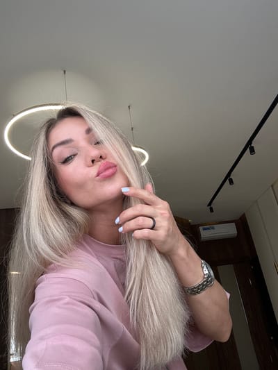 A woman with blonde hair, wearing a pink t-shirt, and a silver watch on her left wrist, is captured in a close-up, selfie-style photograph. Her lips are pursed, and her right hand is raised to her hair, with light blue painted fingernails. She has winged eyeliner and light makeup. The background appears to be an indoor setting, possibly a room, with a ceiling light fixture above and a track lighting system on the ceiling. An air conditioning unit is visible on the wall in the background. The image quality suggests it was taken with a modern smartphone camera. No specific details about the city of Armavir, Russia, are discernible from the visual content of the image.