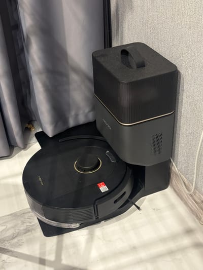 A black Roborock robotic vacuum cleaner is docked with its charging station, which is also black with a gray lower section and a gold accent band. The charging station has a textured top with a black handle. The vacuum cleaner is positioned on a black mat. To the left of the charging station, a grey curtain is partially visible. The background wall has a textured grey pattern, and a white cable runs along the floor and up the wall. The scene appears to be indoors, possibly in a residential setting. The image does not provide any information about the city of Bila Tserkva, Ukraine.