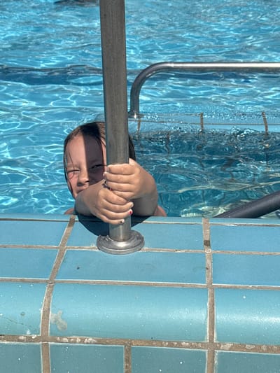 A person is positioned on the edge of a swimming pool, gripping a metal handrail. The pool is tiled with blue tiles, both underwater and on the surrounding deck. The water is clear and reflects sunlight, creating shimmering patterns. The person's hands are visible, with fingernails painted red and black. The person's face is partially submerged, with closed eyes. The location is inferred from prior context to be in Bat Yam, Israel.