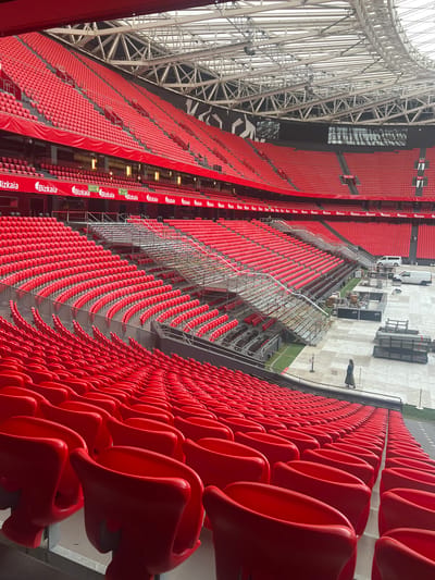 This is a high-angle, wide-shot, exterior photograph of the empty red seats of a large stadium in Bilbao, Spain, viewed from the upper seating tiers. The stadium is predominantly red, with seats stretching across the frame in multiple curved rows. In the background, a white van is parked on a concrete surface, along with stacks of metal pipes. A single person, wearing a long dark coat, is walking on the concrete surface. Above the seats, the stadium's metal roof structure is visible, and a black and white mural or design is painted on the upper portion of the stadium wall. The image appears to have been taken during daylight hours, with the lighting suggesting an overcast sky.