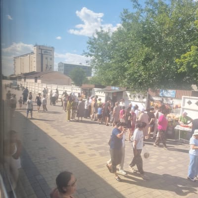 The image shows a busy street scene with a crowd of people gathered near stalls. The location is identified as the city of Turkistan, Kazakhstan. Several buildings are visible in the background, including a large industrial-looking structure on the left and smaller shops or market stalls in the middle ground. The ground is paved with interlocking bricks.
The people in the image are dressed in various casual clothing, with some individuals wearing headscarves. Many people appear to be walking or standing around the stalls, suggesting a market or a gathering place. Some of the stalls are displaying goods, such as food items and drinks. The sky is blue with scattered clouds, and trees are visible on the right side of the image, indicating an outdoor setting on a sunny day. Reflections are visible on the left side of the image, suggesting the photo was taken through a window.