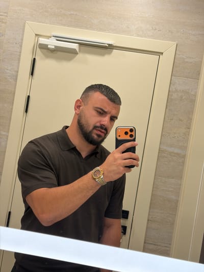 A man with a short haircut and a beard takes a mirror selfie in a bathroom with beige tiled walls and a cream-colored door. He is wearing a dark brown polo shirt and a gold watch with a silver band. He is holding a black smartphone with an orange camera case in his right hand. The phone's screen is not visible. The man's left arm is bent at the elbow, and his left hand is not fully visible. The reflection shows the man from approximately the chest up. The environment appears to be a public or hotel restroom, with a door closer visible at the top of the door frame. There are no other people visible in the image. The lighting is bright, suggesting overhead bathroom lights. The image is taken from a slightly upward angle.