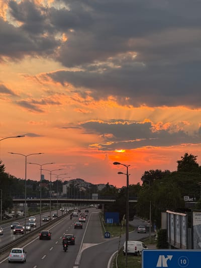 This is a vertical, low-angle, outdoor photograph of a highway during sunset. The sky is filled with dramatic clouds colored orange and yellow by the setting sun, which is partially visible. The highway is busy with traffic, including cars, motorcycles, and a white van. A few streetlights are visible along the side of the road. Trees and buildings line the highway, suggesting an urban or suburban setting. There is a blue road sign with a white arrow indicating a highway exit. The overall mood is serene, with the warm sunset colors creating a peaceful atmosphere. The text "SUPP" is visible on a billboard in the background.