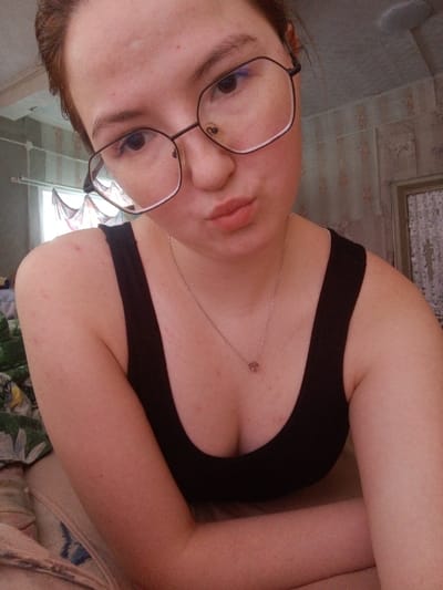 This image captures a head-and-shoulder self-portrait of a young woman in an indoor residential setting in Старый Шагирт, Russia. The light-skinned female individual, with dark hair and dark eyes, is wearing large, dark-framed octagonal glasses. Minor skin blemishes are visible on her forehead, cheeks, and décolletage. Her lips are pursed into a 'kissing' expression. She is dressed in a black sleeveless top with a low neckline, accessorized with a thin silver-colored chain necklace bearing a small, light-colored pendant. Her left arm is bent and partially visible, resting on an unseen surface. The background on the left features a window with a sheer, light-colored, decoratively draped curtain, admitting natural light. In the lower-left, a portion of a patterned fabric, possibly bedding with green and white botanical motifs, is visible. The wall on the right exhibits light-colored, possibly striped wallpaper or paneling. The individual is looking directly at the camera, posing for the photograph.