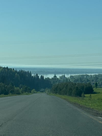 A landscape view of a road curving to the right. To the left of the road, trees and grass grow. Further to the left, a lake or river is visible, and power lines run along the left side of the image. The sky is light blue. The location is Makarovo, Russia.