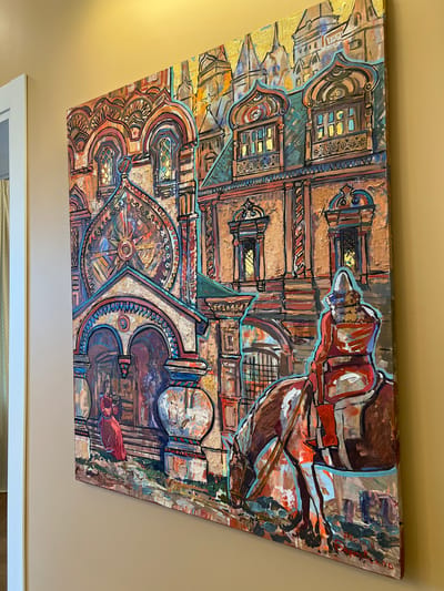 The image is a close-up, angled shot of a vibrantly colored, textured painting mounted on a light beige wall. The painting depicts a stylized architectural scene reminiscent of St. Basil's Cathedral in Moscow, Russia, with its onion domes and ornate facades rendered in bold brushstrokes of reds, oranges, blues, and golds. In the foreground, a figure on horseback is shown from behind, facing away from the viewer. The rider is wearing a red garment, and the horse is a rich brown. To the left, a woman in a red dress stands in an arched entryway of the building. The painting is richly detailed with intricate patterns and decorative elements, suggesting a historical or religious setting. The painting is unsigned, but its style is suggestive of Russian folk art or avant-garde interpretations of traditional Russian architecture. There is no indication of the specific city of Yakunino, Russia, in the image.