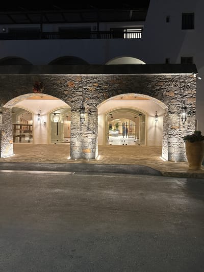 The image depicts the entrance of a building at night, characterized by arched stone structures and illuminated glass doors. A dark asphalt road is in the foreground, leading towards the entrance. The building features multiple arched openings with stone cladding. Decorative lanterns hang from the arches, and subtle ground lighting illuminates the pathway. Inside the building, through the glass doors, a brightly lit interior with modern furnishings and decorative elements is visible. A large potted plant stands to the right of the entrance. The architectural style suggests a modern establishment with traditional elements. The location is identified as Chersonisos Municipal Unit, Greece.