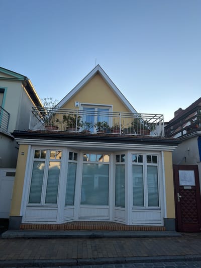 This is a low-angle exterior, eye-level shot of a light yellow building facade under a clear blue sky. The building features a bay window with white mullions and blinds on the ground floor, and a balcony with a metal railing and potted plants on the floor above. The balcony is decorated with several potted plants. The building is flanked by other structures, one to its left with a green roof and another to its right with a dark wooden balcony. The foreground shows a paved sidewalk with brick and stone elements. The scene appears to be located on a street in Rostock, Germany, given the address "48" visible on the building.
