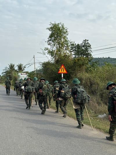 Approximately twelve individuals, identified as soldiers or military trainees, are depicted walking in a column along a paved road in the city of Ninh Hòa, Vietnam. They are viewed from behind, moving away from the camera towards the left of the frame. Each person wears camouflage fatigues, a green pith helmet, and a camouflage backpack. Several individuals have additional gear strapped to their packs, including rolled items and containers, and many carry a long, light-colored wooden staff in their right hand.
The road surface is asphalt, gray in color, and appears to be a two-lane thoroughfare without distinct markings. To the right of the road, dense green and brown vegetation, including bushes and trees, lines the edge. A triangular red and yellow road sign with two vertical black lines (indicating a road narrowing or obstacle) is prominently visible against the vegetation. Power lines are strung between utility poles further in the background on the right. To the left of the road, sparser vegetation and a partially visible large white sign are present. Distant, tree-covered hills or mountains are visible under a cloudy, overcast sky, indicating daylight conditions.