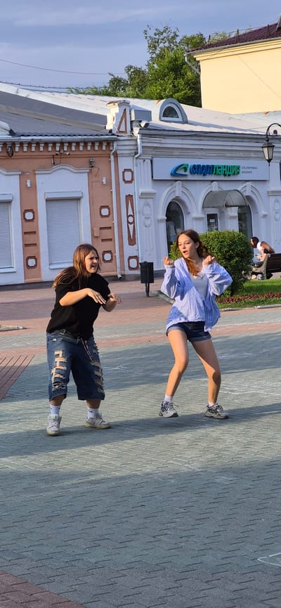 Two young women are dancing on a paved pedestrian area in front of a building with an arched entrance and a sign that reads "Спортландия" (Sportlandia). The woman on the left wears a black t-shirt and distressed denim shorts, while the woman on the right wears a striped blue button-down shirt over a white top and denim shorts. They are positioned mid-action, with their arms and legs extended as if performing a choreographed routine. The background shows a building with decorative architectural elements, trees, and a few other individuals in the distance, one sitting on a bench. The ground is paved with interlocking bricks. The scene appears to be set in an urban environment, likely a public square or street in Ussurijsk, Russia, given the signage. The lighting suggests it is daytime, possibly late afternoon, with sunlight casting shadows on the ground.