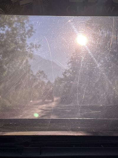 This is a photograph of a view through a scratched window, likely from a vehicle, looking out at a sun-drenched, hazy landscape. The sun, a bright, blown-out circle of light, is prominently featured in the upper right quadrant, casting a glare and creating radial light streaks. The scene outside appears to be a dirt road or path winding through trees, with a blurry mountain range visible in the distance. The lower portion of the image is dominated by the dark, textured frame of the window or vehicle interior. There is a faint green circular artifact visible in the lower left quadrant, possibly a lens flare or reflection. The image is characterized by numerous scratches and smudges on the window surface, obscuring the details of the outdoor scene. The location in Kintamani, Indonesia, is not discernible from the visual content of the photograph itself.